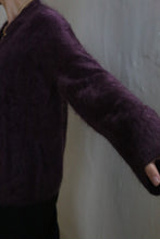 Load image into Gallery viewer, A person in the Brushed Cashmere Cardigan in Aubergine poses against a light-colored textured wall, showcasing only the upper body and outstretched arm.