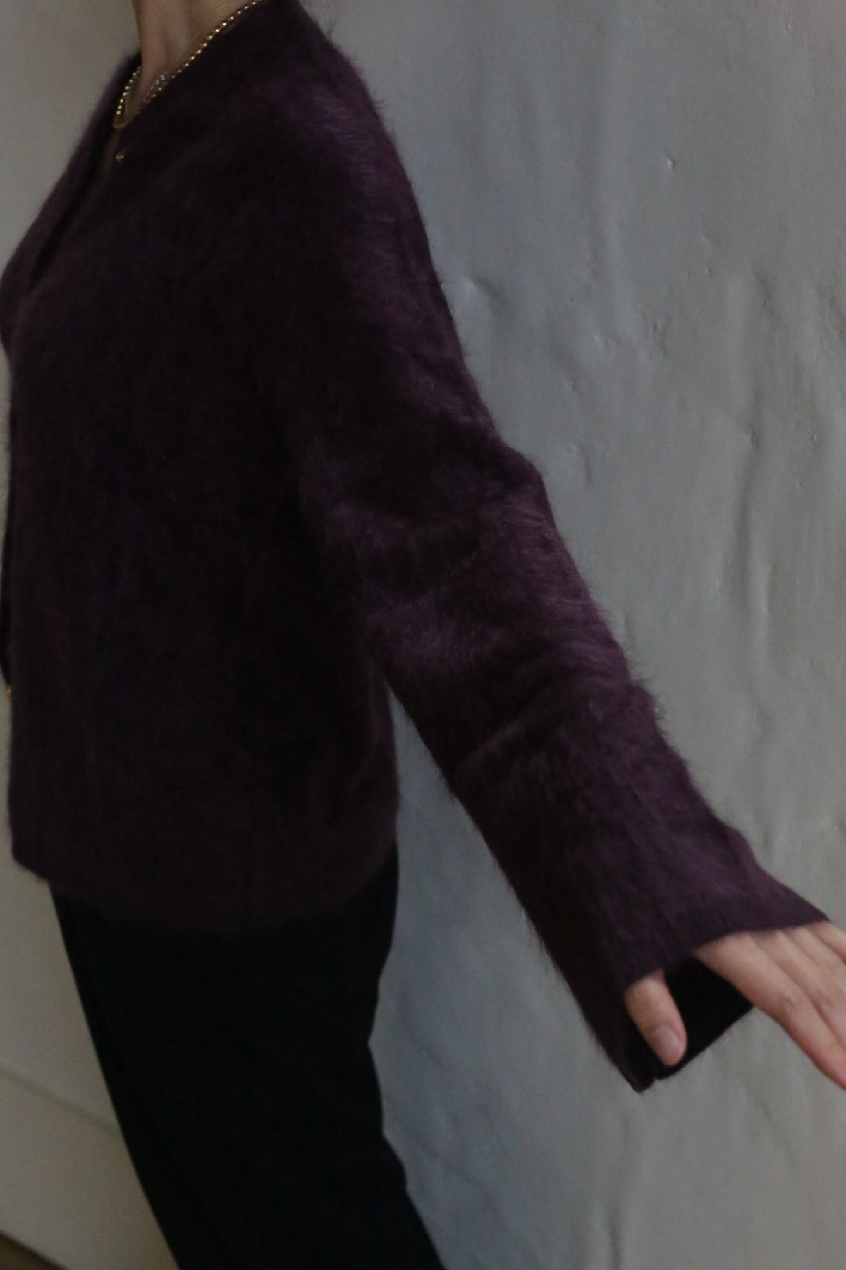 A person models the Brushed Cashmere Cardigan in Aubergine, featuring long flared sleeves and a cozy, luxurious knit, paired with black pants against a plain light wall.