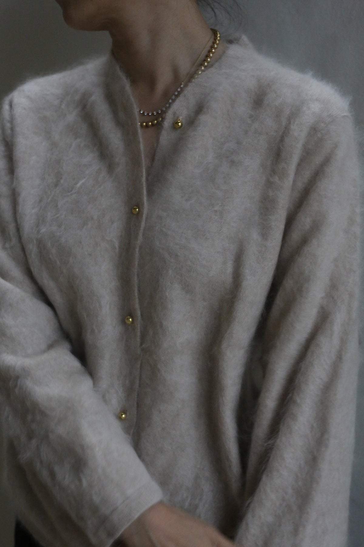 A person wears the Brushed Cashmere Cardigan in Sandstone with brass buttons and a gold beaded necklace, standing against a neutral background. Their face is partially out of frame.