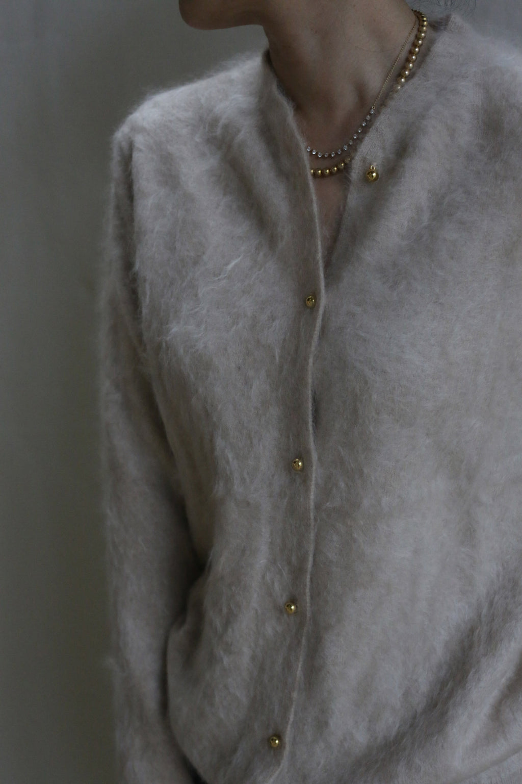 A person wearing a soft Brushed Cashmere Cardigan in Sandstone with gold buttons and layered gold necklaces; only the upper torso and part of the neck are visible.