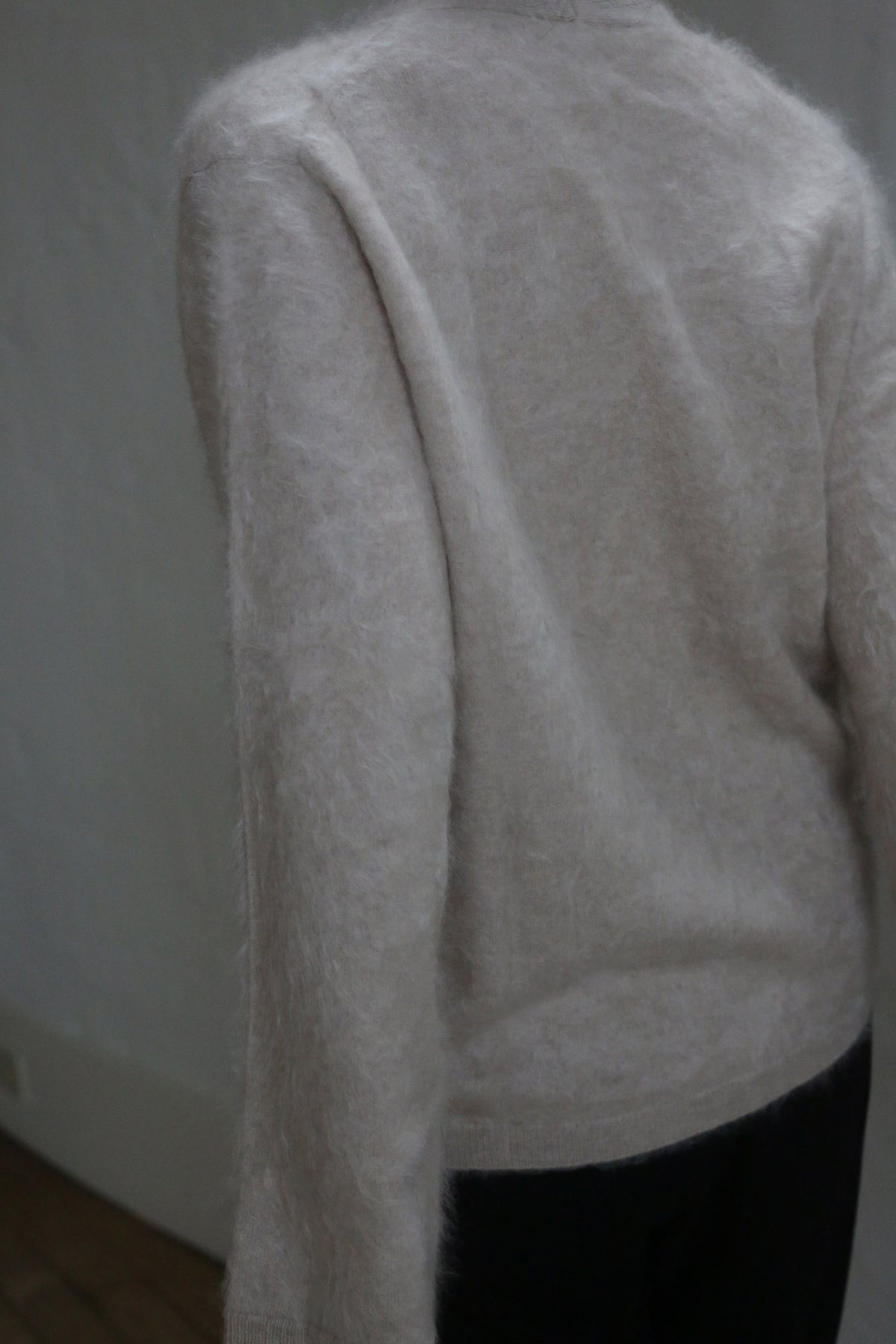 Someone stands with their back to the camera, one arm slightly bent, wearing a soft, light Brushed Cashmere Cardigan in Sandstone with brass buttons. The softly lit background is neutral.
