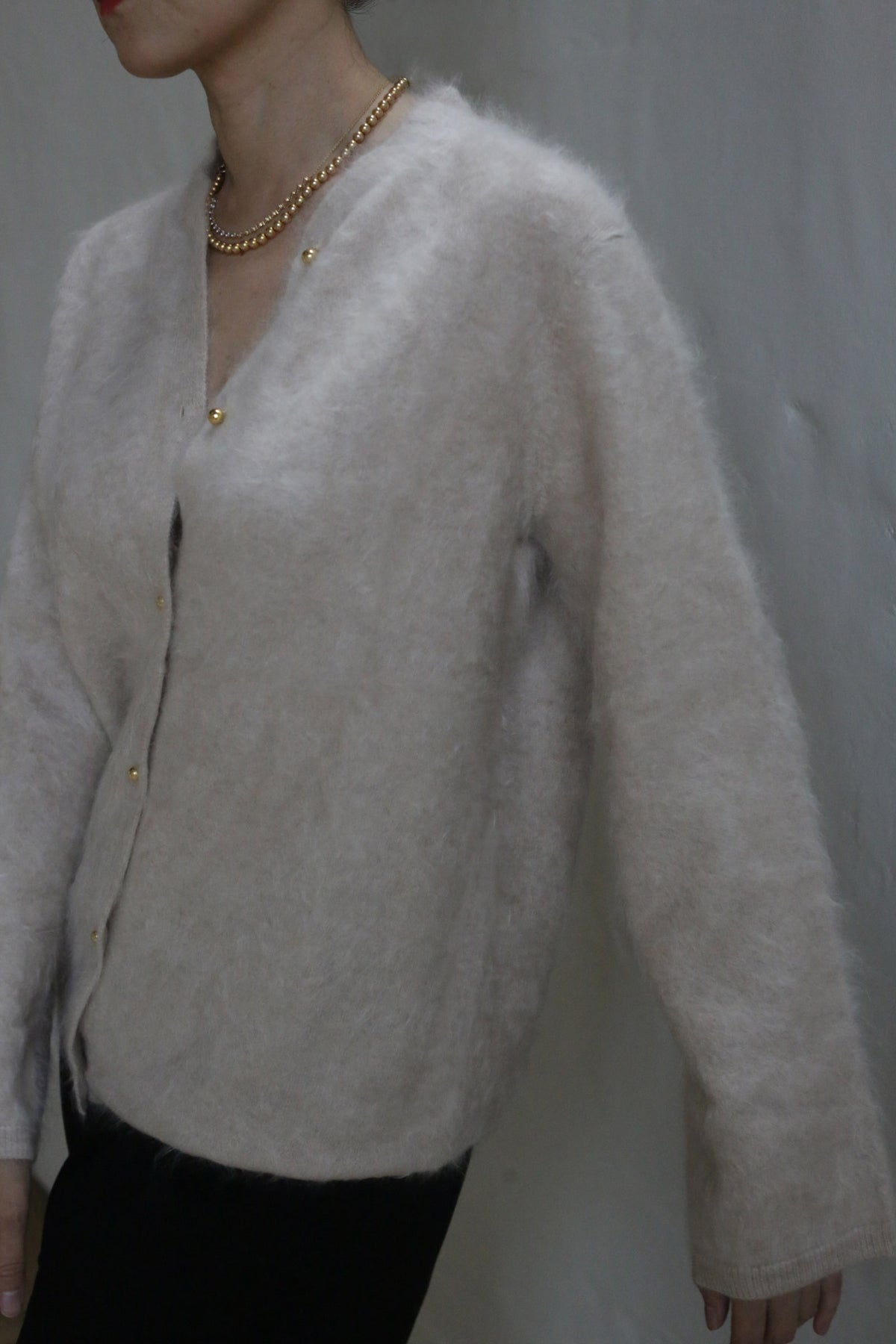 A person wearing the Brushed Cashmere Cardigan in Sandstone with brass buttons and a gold chain necklace stands against a plain light-colored wall, their face partially out of frame.