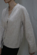 Load image into Gallery viewer, A person wearing the Brushed Cashmere Cardigan in Sandstone with brass buttons and a gold chain necklace stands against a plain light-colored wall, their face partially out of frame.
