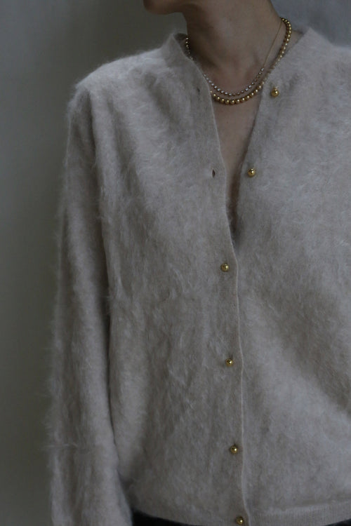 A person models the Brushed Cashmere Cardigan in Sandstone, featuring brass buttons and layered gold necklaces, photographed from the shoulders down against a neutral backdrop.
