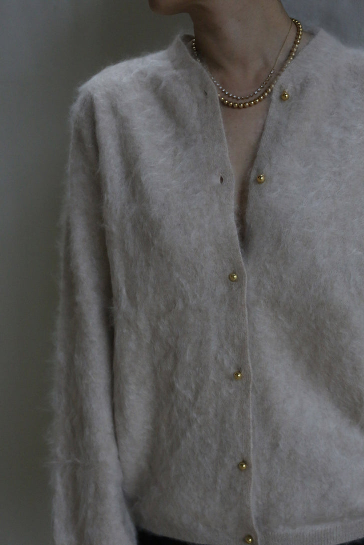 A person models the Brushed Cashmere Cardigan in Sandstone, featuring brass buttons and layered gold necklaces, photographed from the shoulders down against a neutral backdrop.