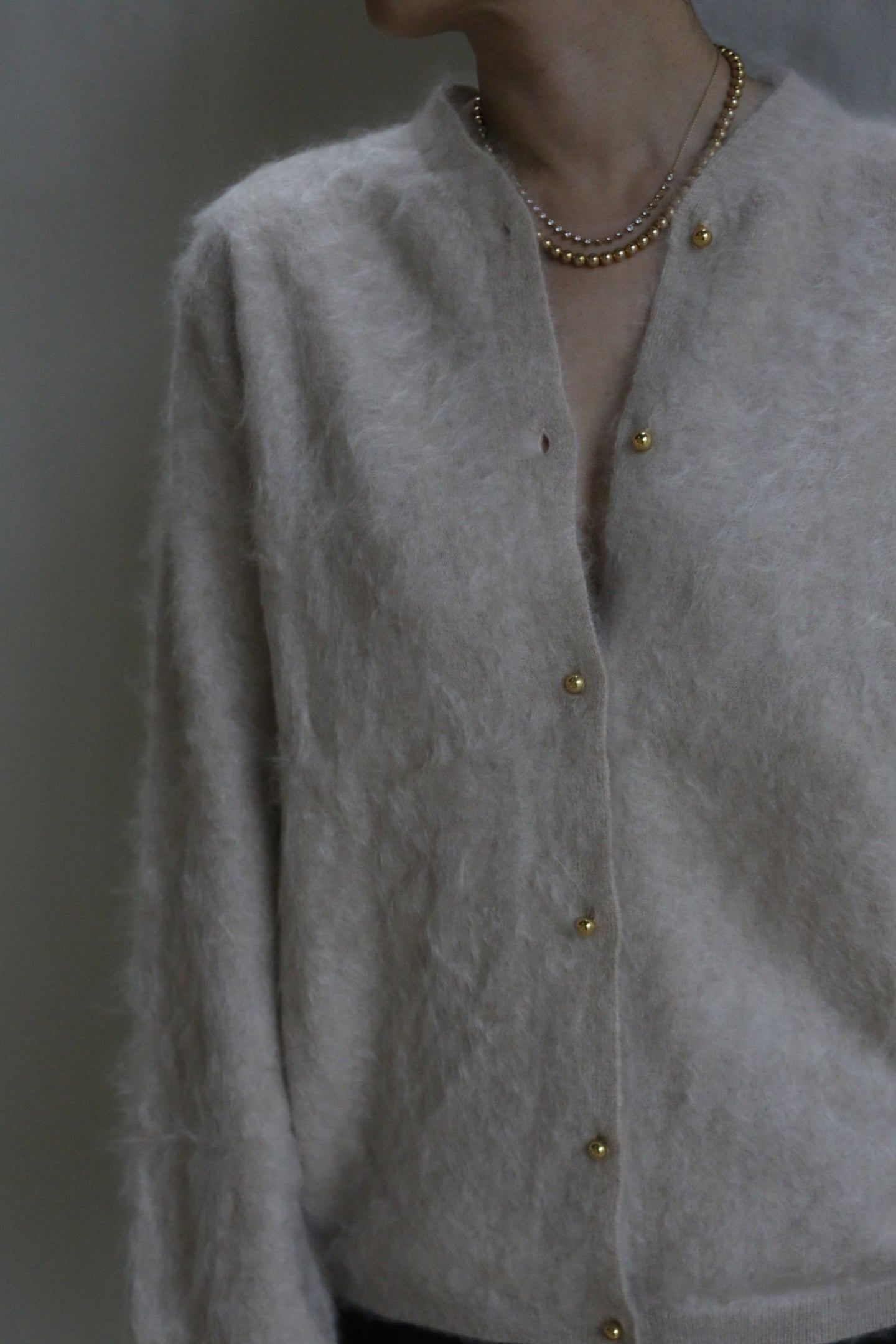 A person models the Brushed Cashmere Cardigan in Sandstone, featuring brass buttons and layered gold necklaces, photographed from the shoulders down against a neutral backdrop.