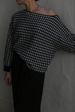 Load image into Gallery viewer, The individual is styled in the Ballerina&#39;s Cashmere Sweater | Houndstooth Jacquard, featuring a black and white houndstooth pattern and an off-the-shoulder boat neck design. They accessorize with a gold beaded necklace while posing against a plain background, with their face partially obscured.