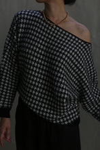 Load image into Gallery viewer, An individual is dressed in the Ballerina&#39;s Cashmere Sweater with a houndstooth jacquard pattern, featuring an off-the-shoulder design and a relaxed fit, paired with a gold beaded necklace, standing against a gray background.