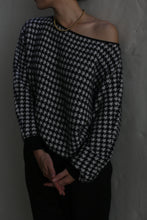 Load image into Gallery viewer, A person wearing a Ballerina&#39;s Cashmere Sweater with a relaxed fit and houndstooth jacquard pattern, paired with a gold beaded necklace, stands against a plain wall. Their hands are folded in front of them, and their face is turned away from the camera.