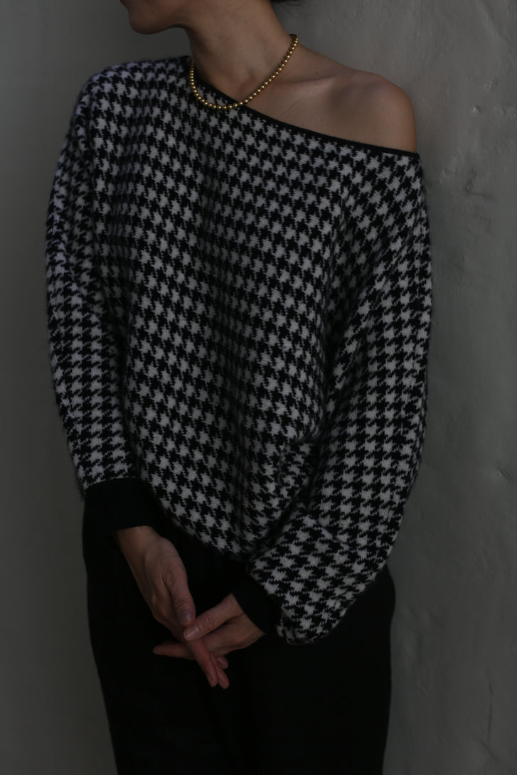 A person wearing a Ballerina's Cashmere Sweater with a relaxed fit and houndstooth jacquard pattern, paired with a gold beaded necklace, stands against a plain wall. Their hands are folded in front of them, and their face is turned away from the camera.