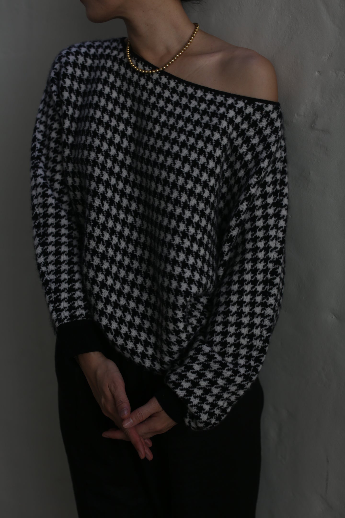 A person wearing a Ballerina's Cashmere Sweater with a relaxed fit and houndstooth jacquard pattern, paired with a gold beaded necklace, stands against a plain wall. Their hands are folded in front of them, and their face is turned away from the camera.