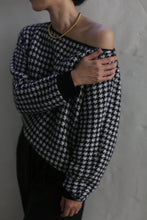 Load image into Gallery viewer, A person wearing the Ballerina&#39;s Cashmere Sweater in a houndstooth jacquard design and a gold beaded necklace stands against a textured wall, with one hand resting on their opposite shoulder, partially covering their face.