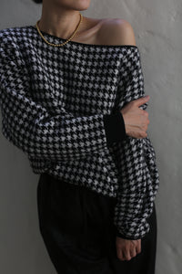 A person wearing the Ballerina's Cashmere Sweater with a houndstooth jacquard pattern in an off-shoulder, relaxed fit style, complemented by a gold necklace, stands against a neutral background with arms crossed.