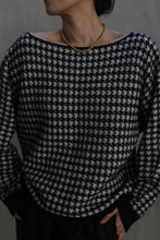 Load image into Gallery viewer, A person wearing the Ballerina&#39;s Cashmere Sweater in houndstooth jacquard, featuring a relaxed fit and wide neckline, is adorned with a gold beaded necklace. Their hands rest in their pants pockets as they turn their face slightly away from the camera.