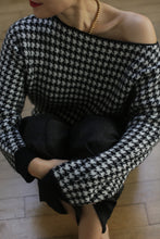 Load image into Gallery viewer, Person wearing the Ballerina&#39;s Cashmere Sweater in a black and white houndstooth jacquard design, featuring a relaxed fit. They pair it with dark pants and a gold necklace while sitting on a wooden floor with arms crossed over their knees, their face slightly turned away revealing red lipstick.