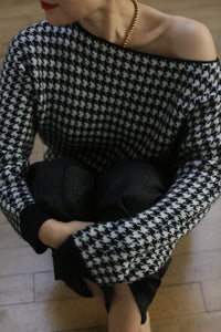 Person wearing the Ballerina's Cashmere Sweater in a black and white houndstooth jacquard design, featuring a relaxed fit. They pair it with dark pants and a gold necklace while sitting on a wooden floor with arms crossed over their knees, their face slightly turned away revealing red lipstick.