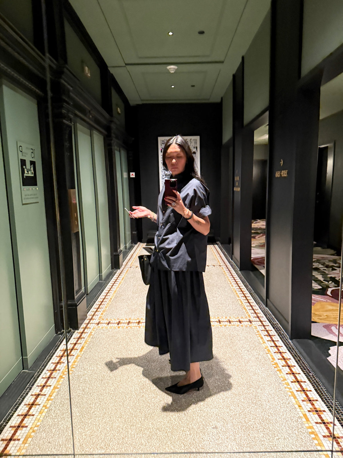 Wearing the NYFW Edition — Work Shirt & Wrap Skirt Set and black heels, a person takes a mirror selfie in a hotel hallway with green walls, patterned carpet, and several elevator doors.