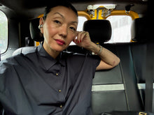 Load image into Gallery viewer, A woman with dark hair pulled back sits in the back seat of a car, wearing the Work Shirt | Typewriter Cotton Black, accessorized with jewelry, looking at the camera with her head resting on her hand. The car interior and window are visible behind her.