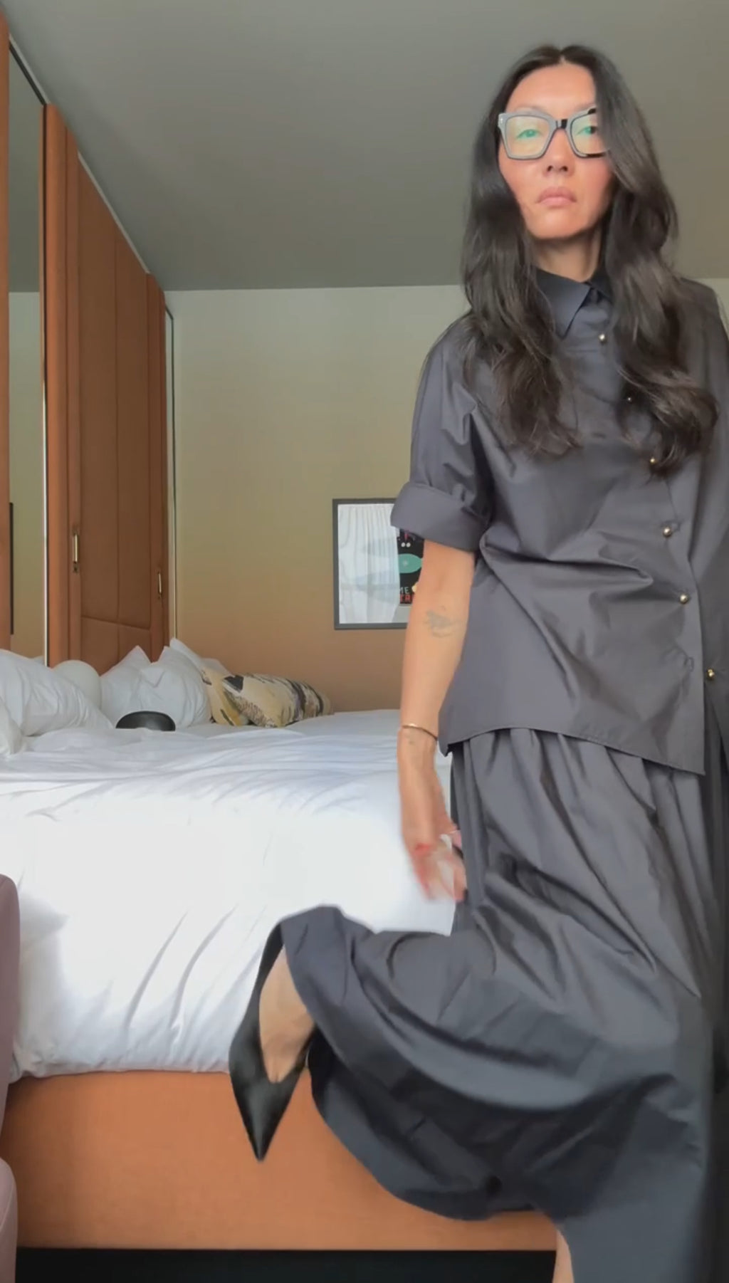 A woman with long dark hair and glasses models the NYFW Edition — Work Shirt & Wrap Skirt Set in dark gray, posing in a bedroom with an unmade bed and lifting her skirt slightly to reveal black shoes.