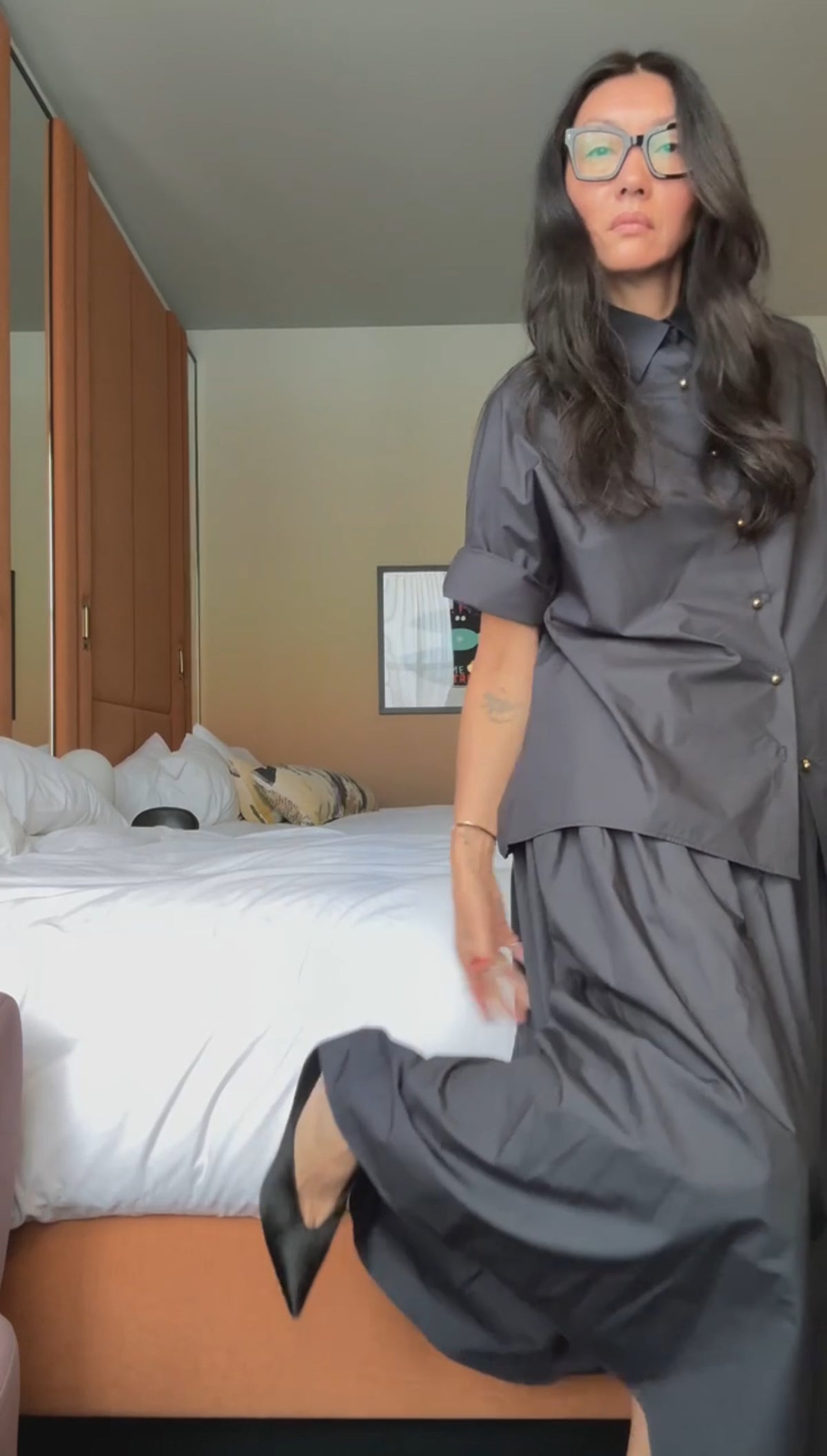 A woman with long dark hair and glasses models the NYFW Edition — Work Shirt & Wrap Skirt Set in dark gray, posing in a bedroom with an unmade bed and lifting her skirt slightly to reveal black shoes.
