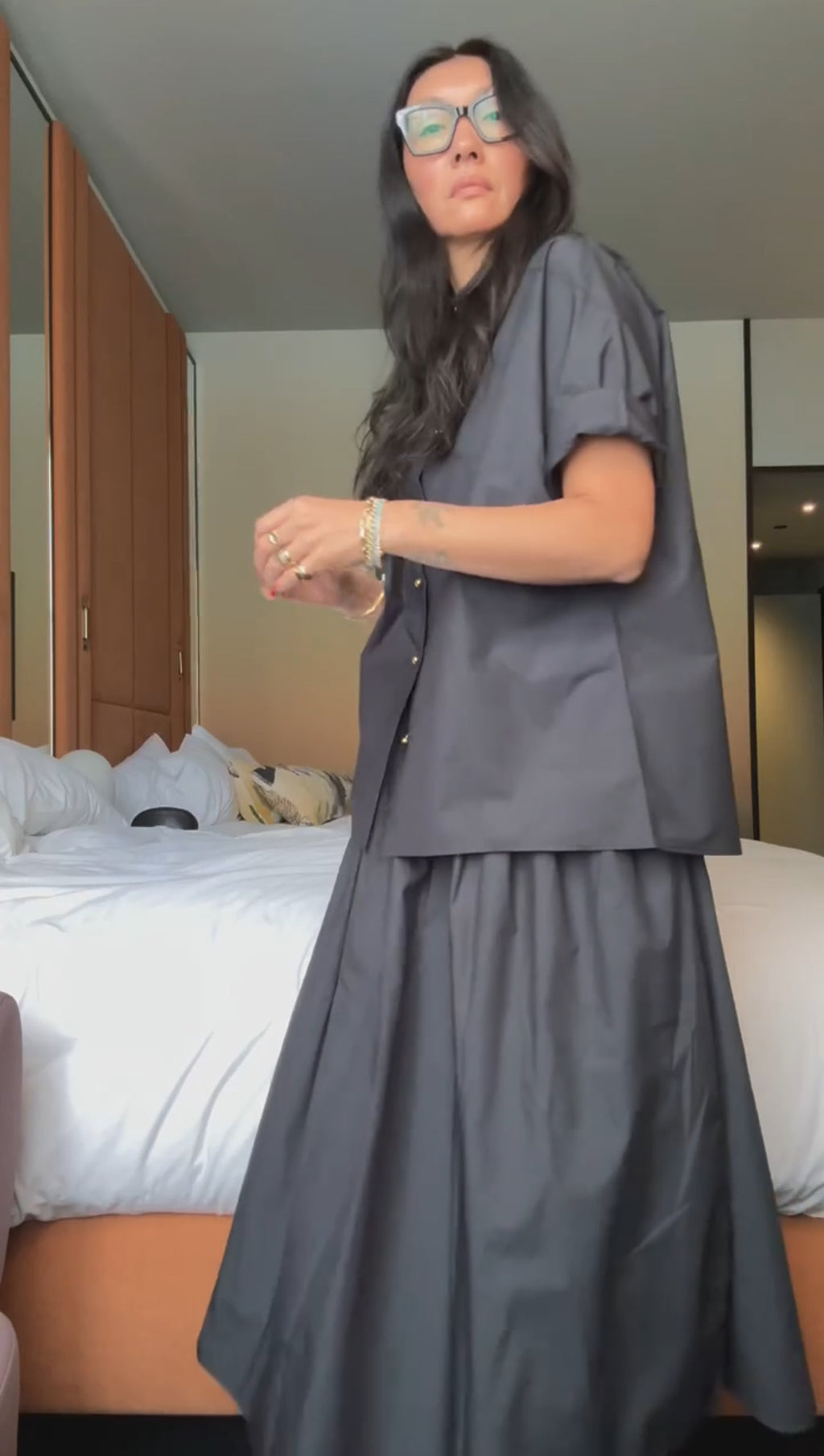 A woman with long dark hair and glasses stands indoors in the NYFW Edition — Work Shirt & Wrap Skirt Set, with a made bed and wooden cabinets in the background.