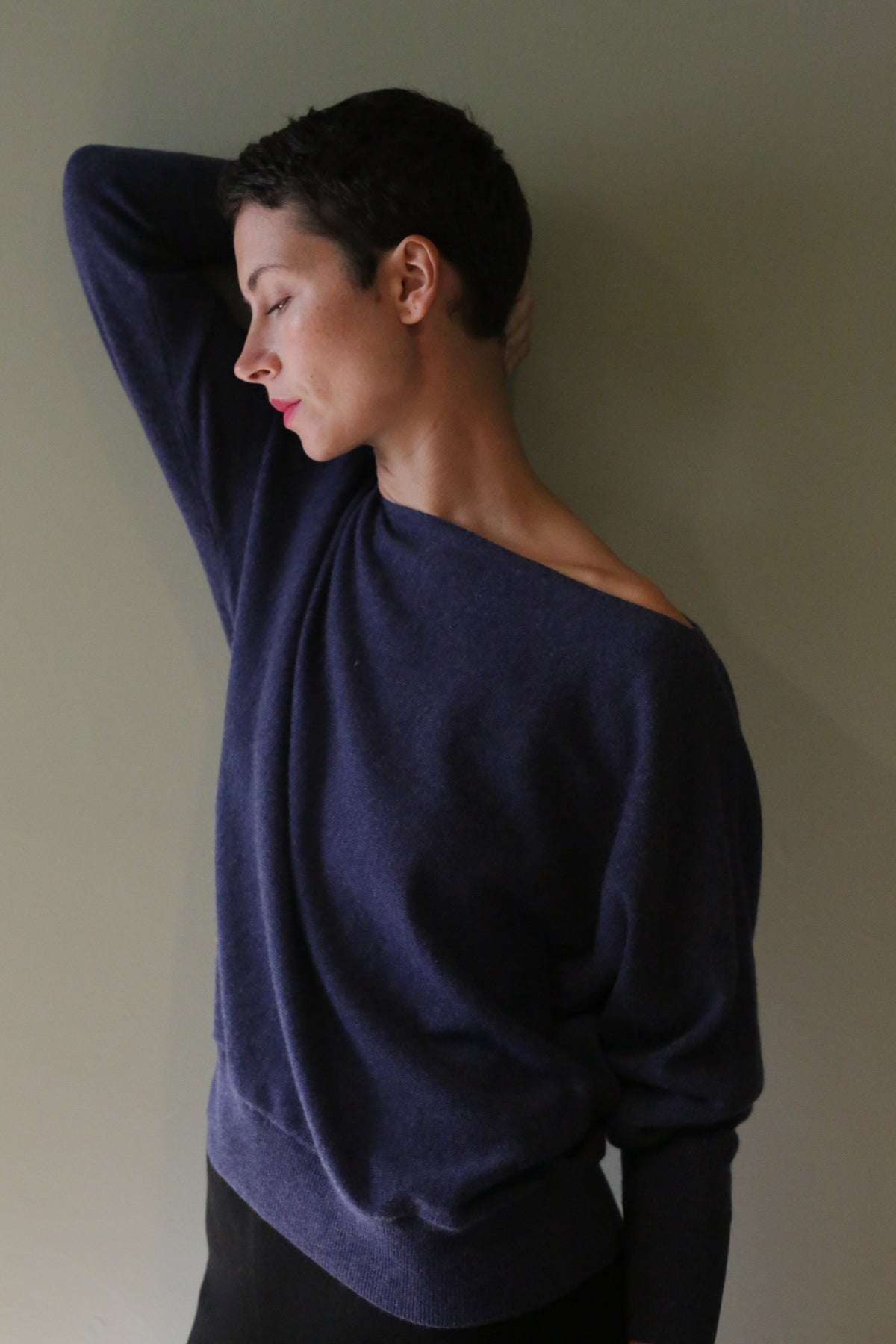 A person with short hair wears a navy blue, off-the-shoulder long-sleeved Ballerina’s Cashmere Sweater | Navy in a relaxed silhouette, with one arm raised.