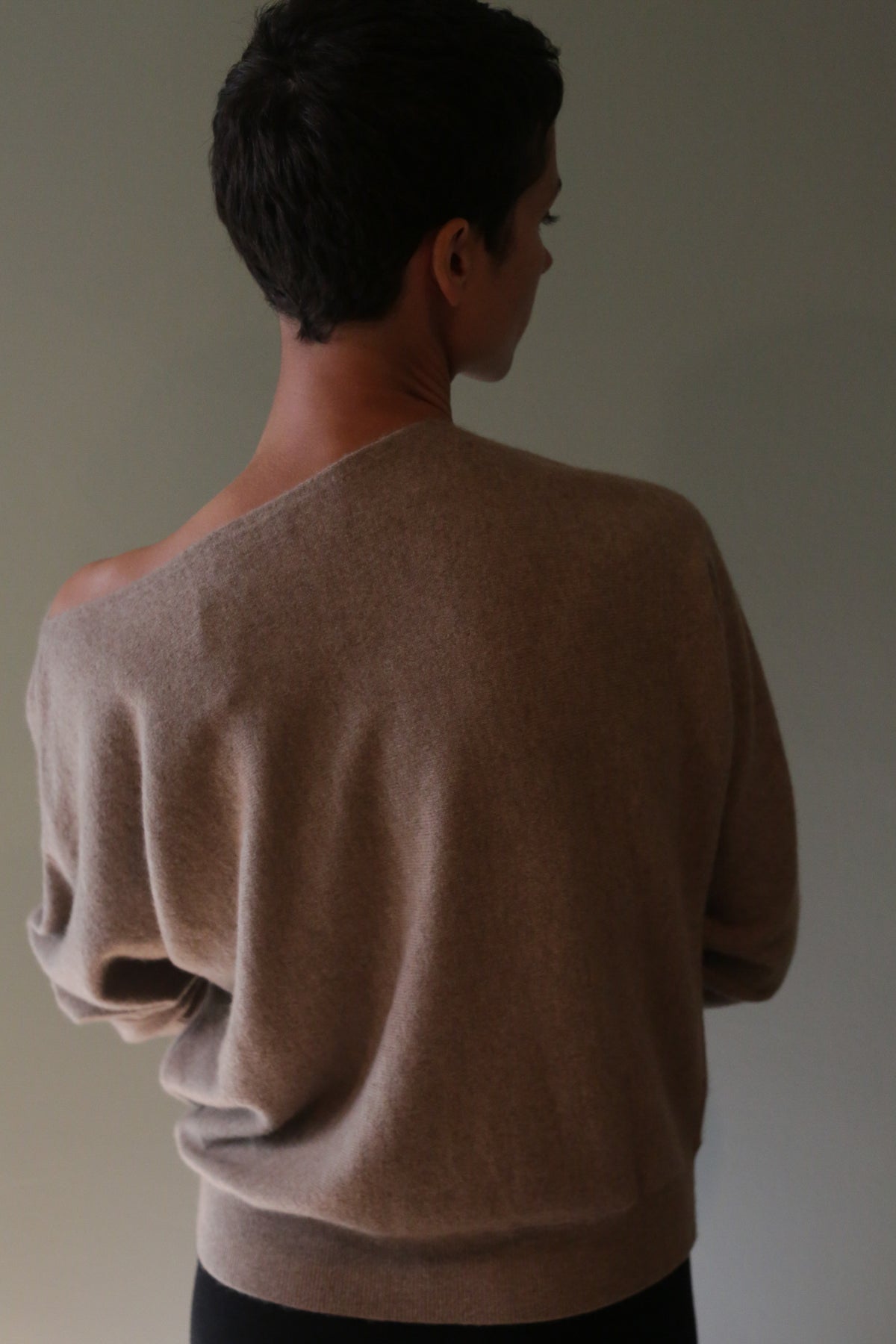 A person wearing the Ballerina’s Cashmere Sweater | Oat, a relaxed, oat-colored cashmere sweater with an asymmetrical neckline that slips off one shoulder.