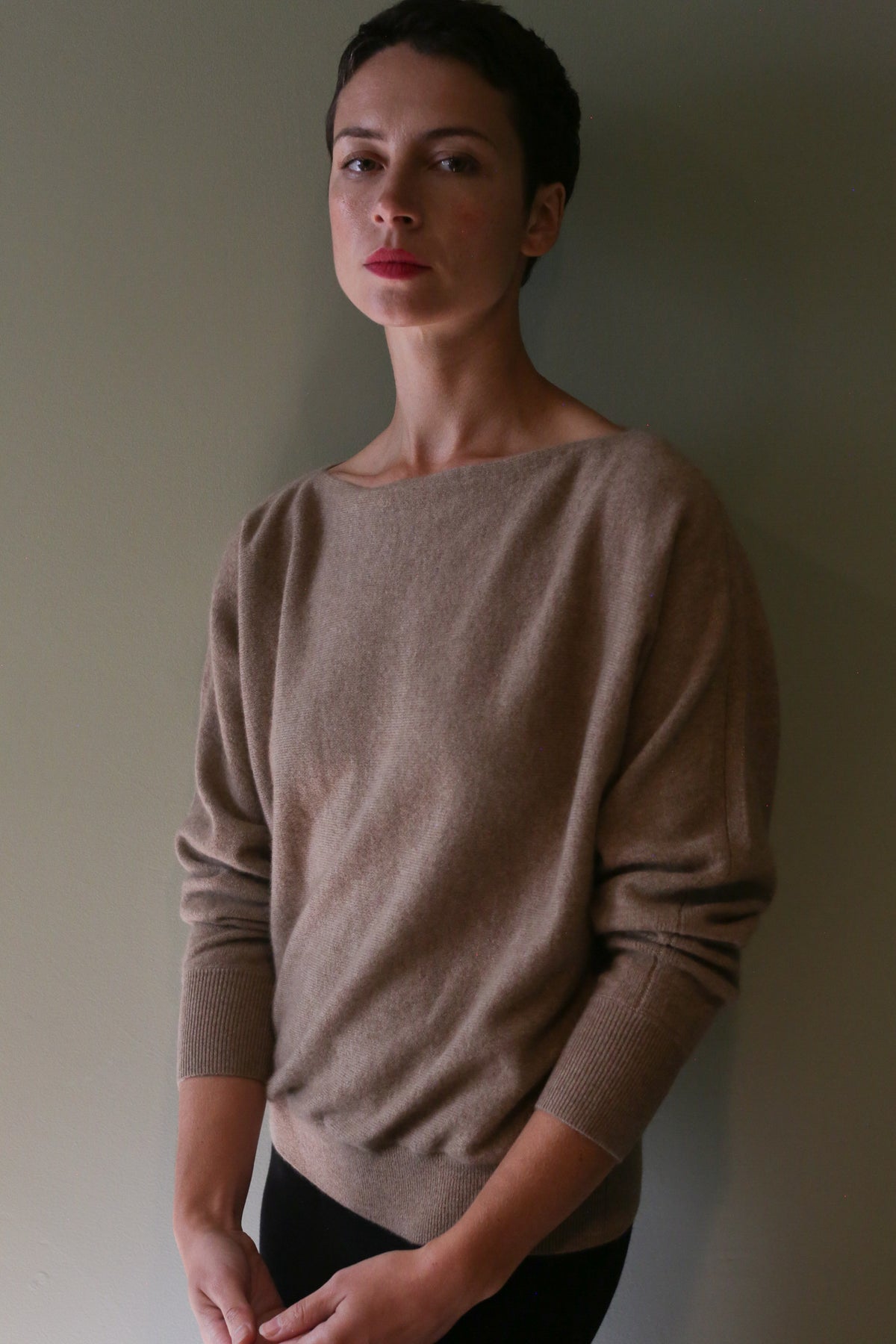 A person wearing the Ballerina’s Cashmere Sweater | Oat, a beige cashmere boat-neck sweater with long sleeves and ribbed cuffs in a relaxed silhouette.