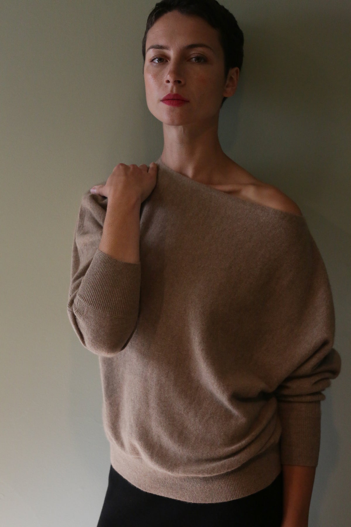 A person with short dark hair wears an off-the-shoulder Ballerina’s Cashmere Sweater in oat, featuring a tan hue and a relaxed silhouette.
