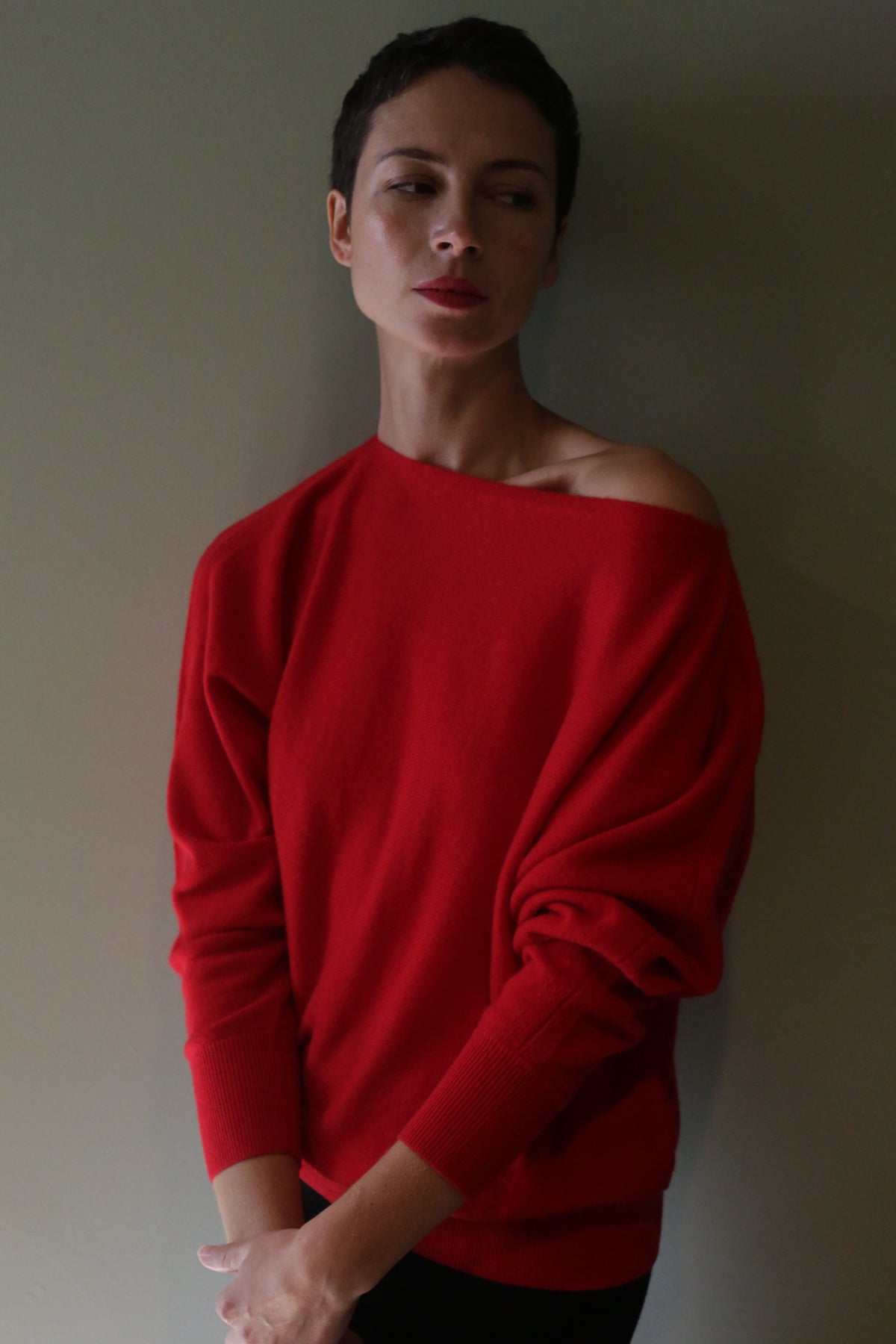A person wears a relaxed-silhouette Ballerina’s Cashmere Sweater in vermillion with an asymmetrical neckline that drops off one shoulder.
