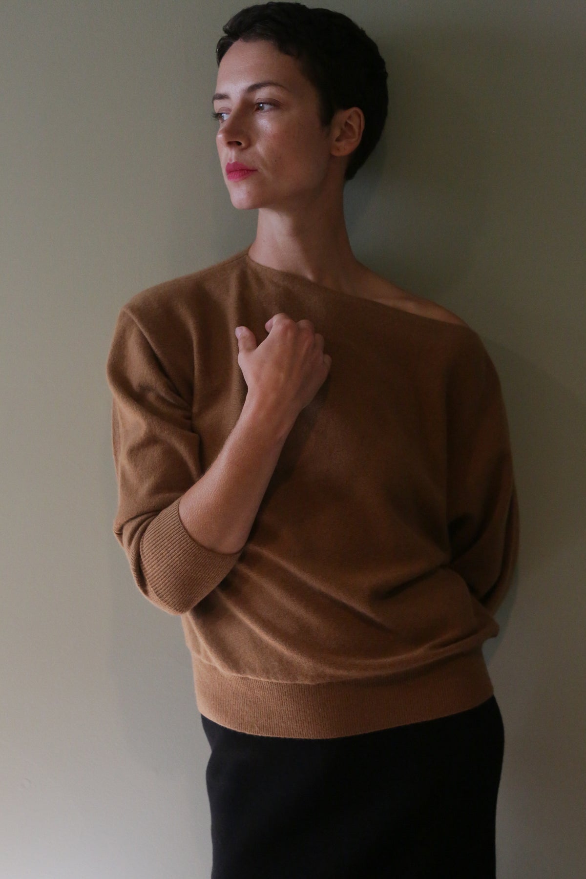 A person wearing a Ballerina’s Cashmere Sweater in Vicuna, featuring a brown, long-sleeved, relaxed-fit design with an asymmetrical neckline.