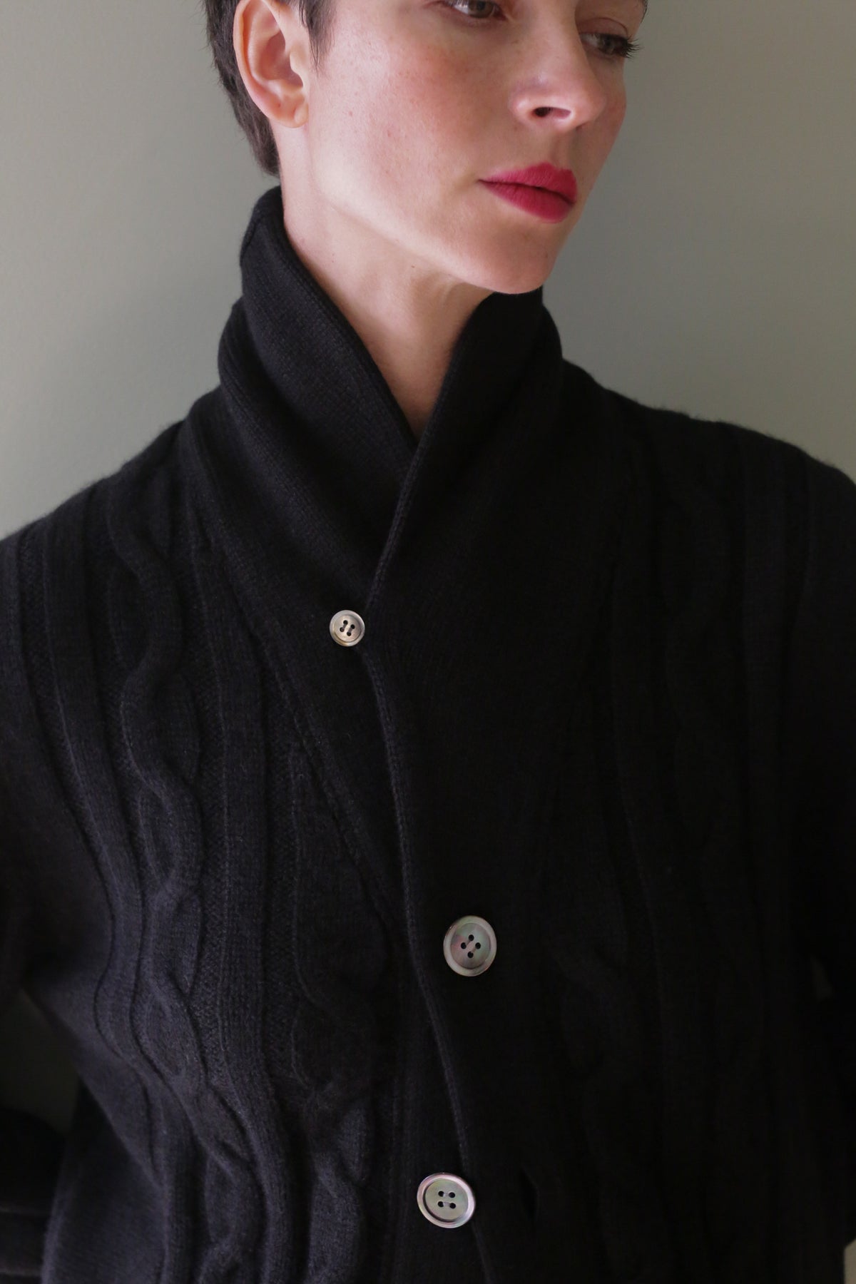 A person wears a black Unisex Shawl-Collar Cable-Knit Cashmere Cardigan featuring a shawl collar and three buttons.