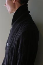 Load image into Gallery viewer, Profile view of a person wearing a black Unisex Shawl-Collar Cable-Knit Cashmere Cardigan with a wide shawl collar and visible buttons.