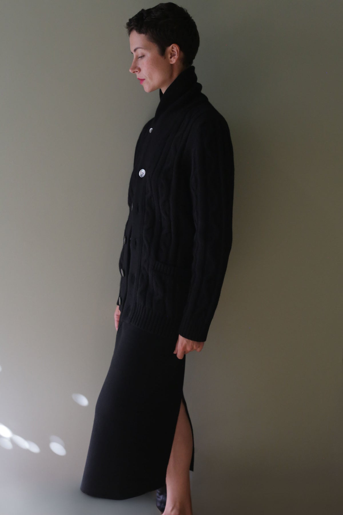 Profile view of a person wearing a black Unisex Shawl-Collar Cable-Knit Cashmere Cardigan | Black with large buttons over a matching black skirt.