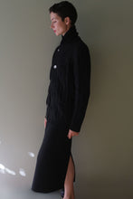 Load image into Gallery viewer, Profile view of a person wearing a black Unisex Shawl-Collar Cable-Knit Cashmere Cardigan | Black with large buttons over a matching black skirt.