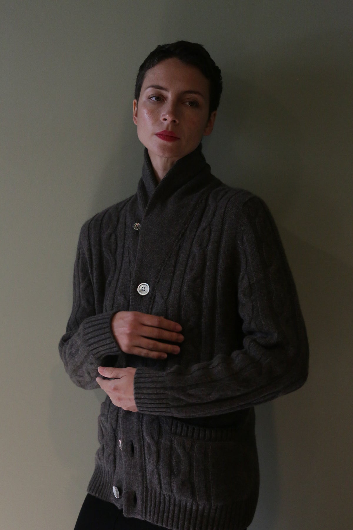 A person wearing a Unisex Shawl-Collar Cable-Knit Cashmere Cardigan | Taupe, a dark brown shawl-collar cashmere cardigan with a cable-knit texture and large white buttons.
