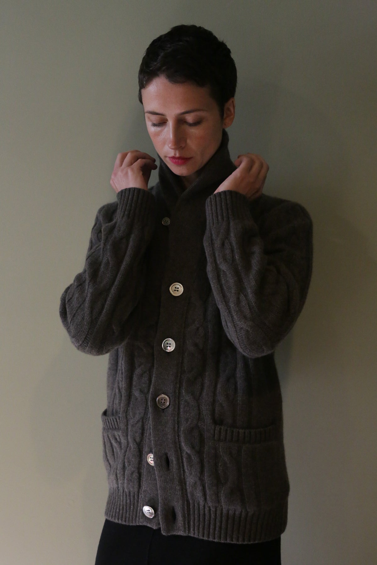 A person holds the lapels of a taupe Unisex Shawl-Collar Cable-Knit Cashmere Cardigan, featuring a shawl collar, button closure, and cable-knit texture.