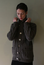 Load image into Gallery viewer, A person holds the lapels of a taupe Unisex Shawl-Collar Cable-Knit Cashmere Cardigan, featuring a shawl collar, button closure, and cable-knit texture.