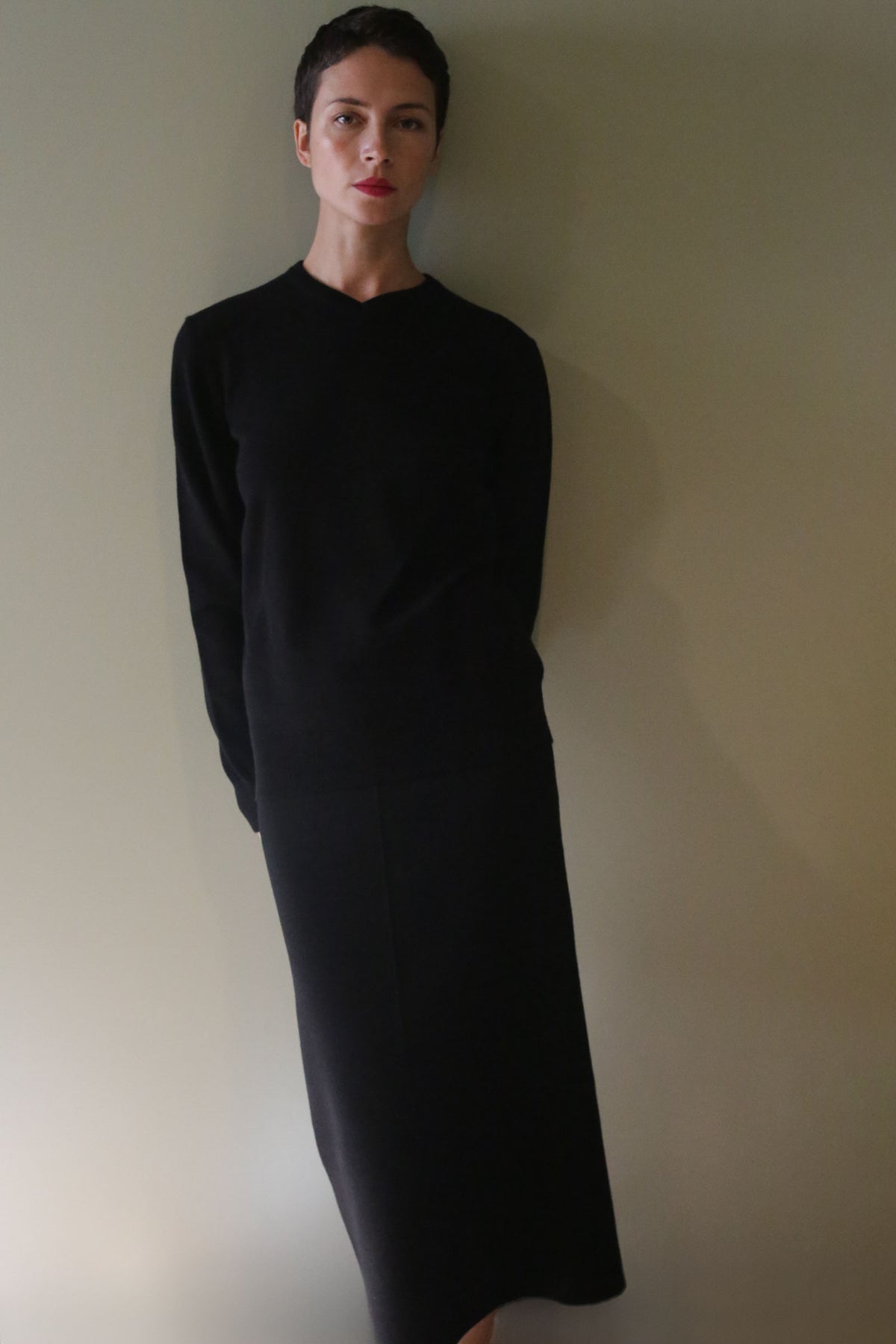 A person wearing a black long-sleeved top and a matching Cashmere Pencil Skirt | Black standing against a plain wall.