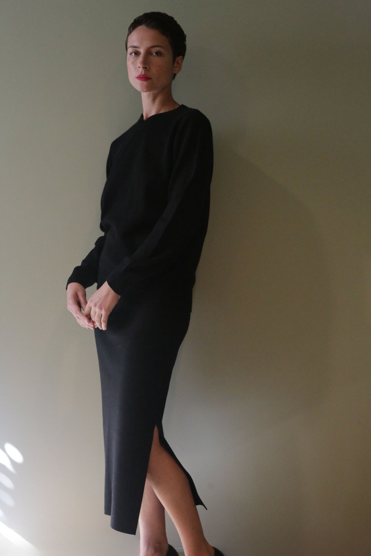 A person wearing a black long-sleeved sweater and a matching Cashmere Pencil Skirt | Black featuring a side slit.