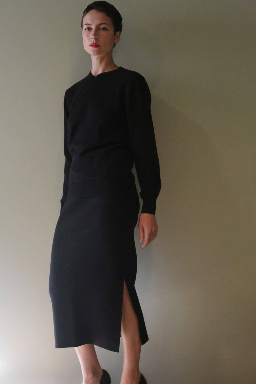 A person wearing a long-sleeved black top and a matching Cashmere Pencil Skirt | Black featuring an individually knitted design and a side slit.