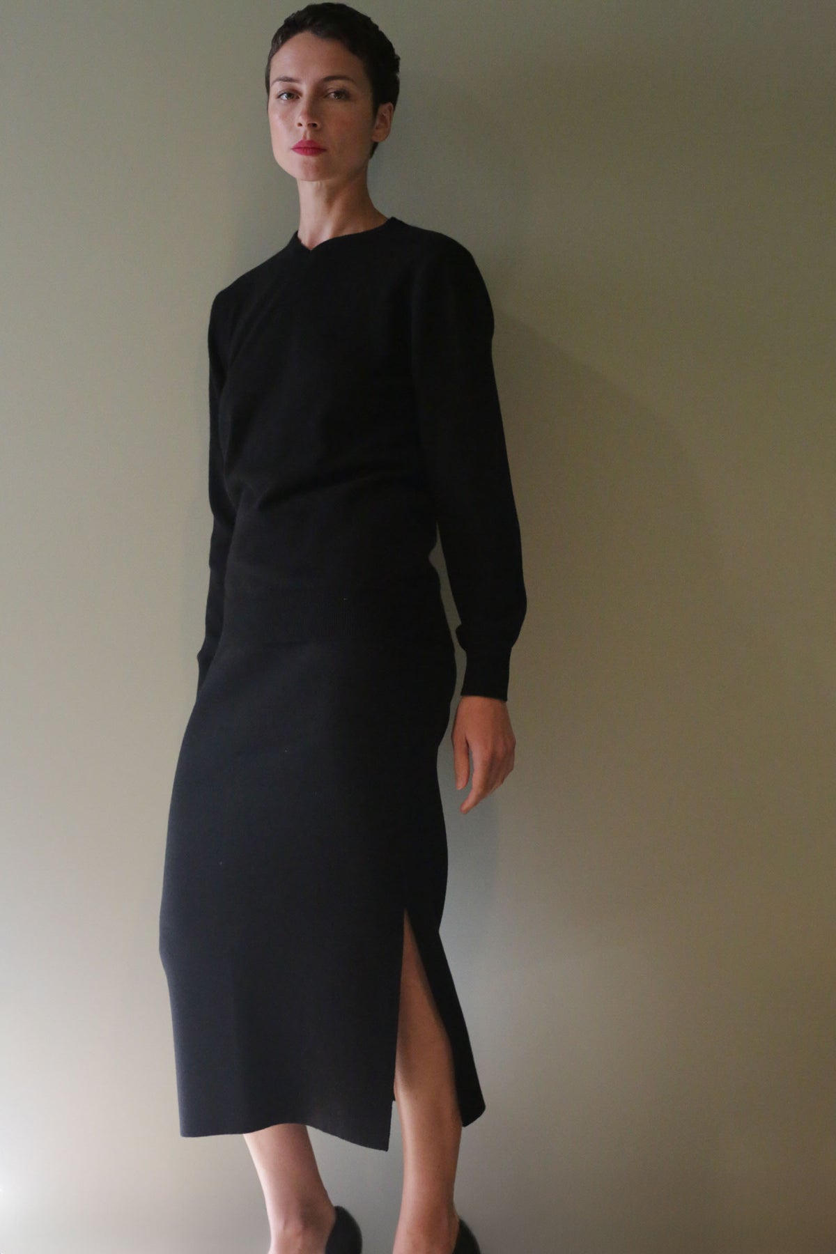A person wearing a long-sleeved black top and a matching Cashmere Pencil Skirt | Black featuring an individually knitted design and a side slit.