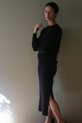 A person wearing a black long-sleeved top and a matching Cashmere Pencil Skirt | Black featuring a side slit.