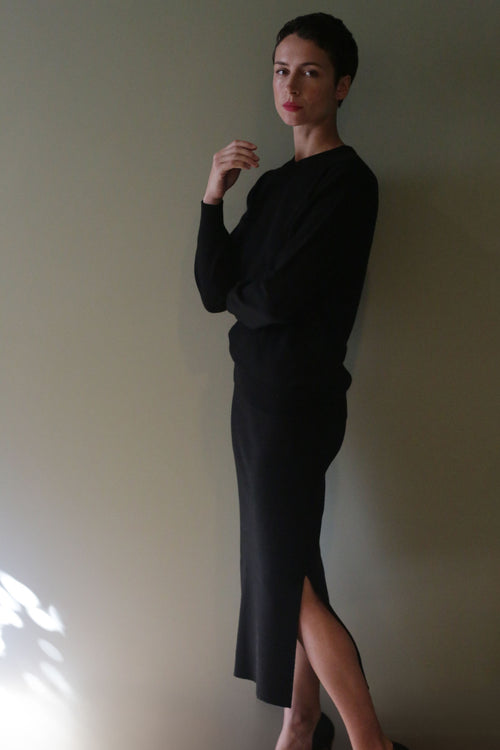 A person wearing a black long-sleeved top and a matching Cashmere Pencil Skirt | Black featuring a side slit.