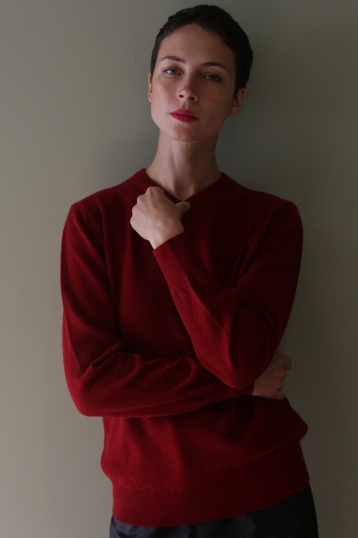 Person with short dark hair wears the High V-Neck Cashmere Sweater | Burgundy, standing confidently against a plain light wall with one arm crossed and the other hand touching their neck.