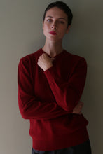 Load image into Gallery viewer, Person with short dark hair wears the High V-Neck Cashmere Sweater | Burgundy, standing confidently against a plain light wall with one arm crossed and the other hand touching their neck.