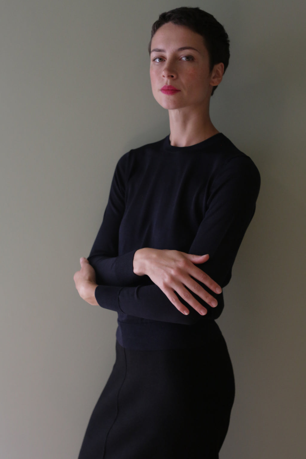 A woman with short dark hair wears a black Washable Silk Crewneck, an individually knitted, long-sleeved sweater, with her arms crossed.