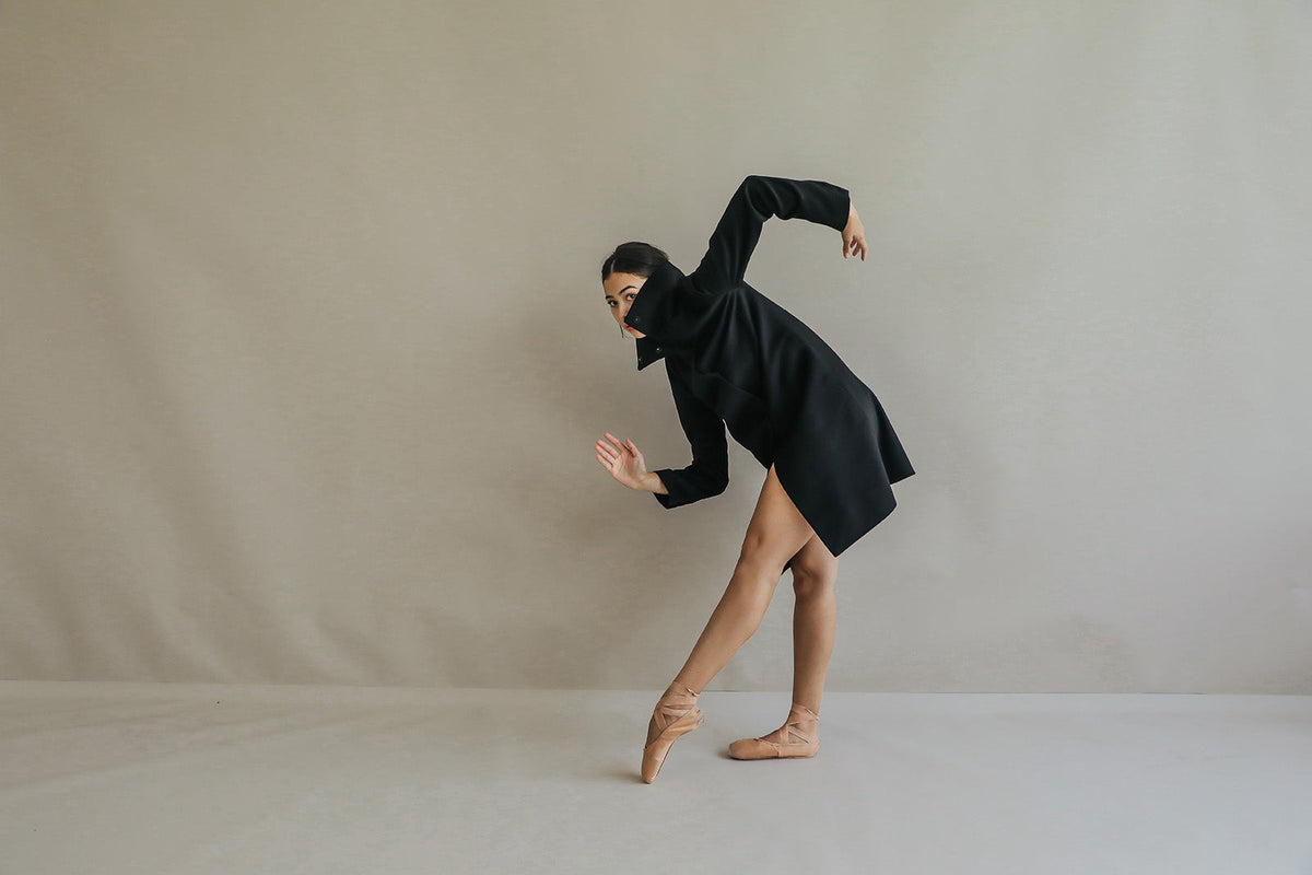A person in a black Stamford Long | Black straight-cut cashmere coat strikes a ballet pose with one hand raised to their face.