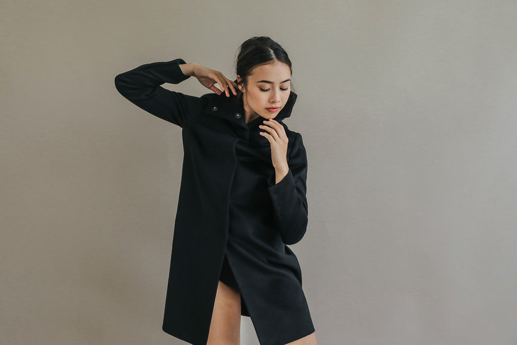 A woman wearing the black, straight-cut Stamford Long | Black cashmere coat with a high collar, posing with her hand near her neck.