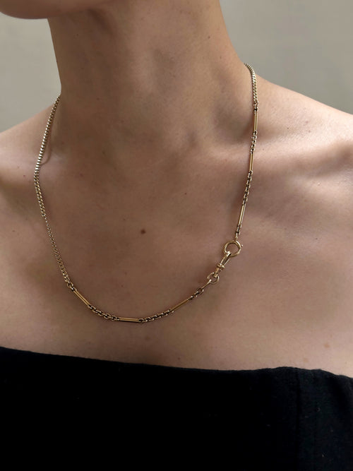 A person wearing an off-the-shoulder black top and an Antique 14K Gold Bar and Link Chain c.1900 with a detailed clasp, complemented by a 12K Flat Curb Chain c.1880. The necklace features a combination of elongated and grooved bar links alongside smaller circular links, set against a plain, neutral background.
